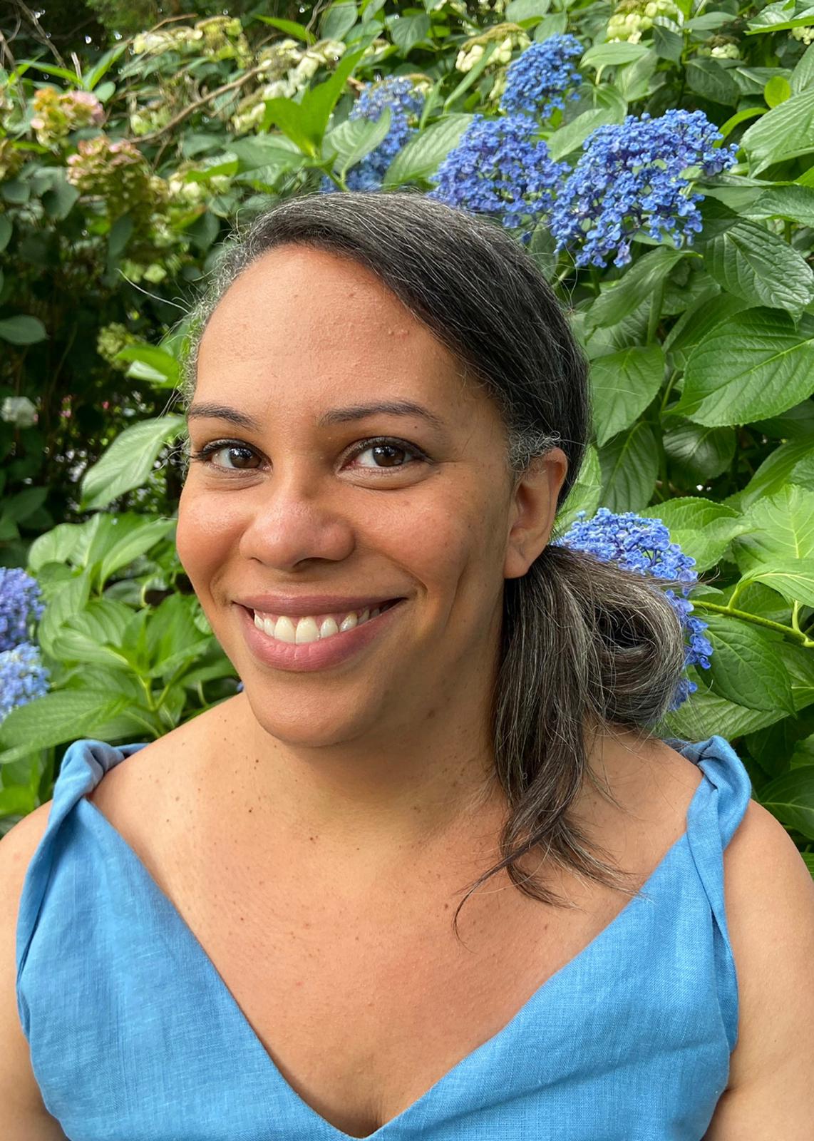 A close up portrait of Amara Thornton. She is smiling in front of a flowering Hydrangea bush.