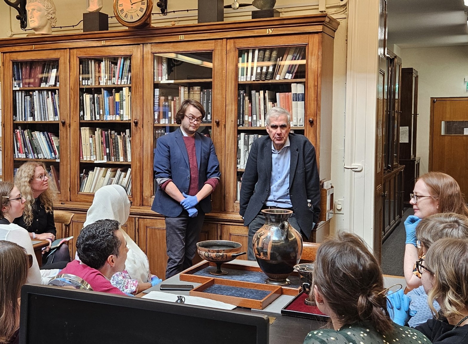 In the British Museum Antiquities reading room, a small group of people are seated round a table with three Greek Pots arranged in low trays. There are two speakers behind the pots. The older man (Thomas Mannack) leans forward to add emphasis to a point and his colleague (Ollie Croker) raises an eyebrow. Everybody is wearing blue gloves to handle the pots.
